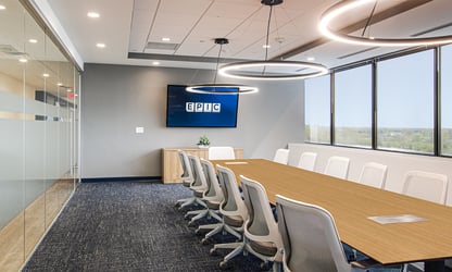 Modern conference room with a long table, white chairs, large windows, and a screen displaying 