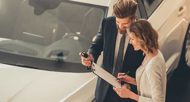 an automotive sales associate discusses a contract with a customer while standing next to a sedan