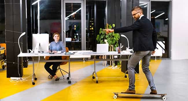 a real estate executive rides a skateboard through an office lobby past a colleague working at a desk