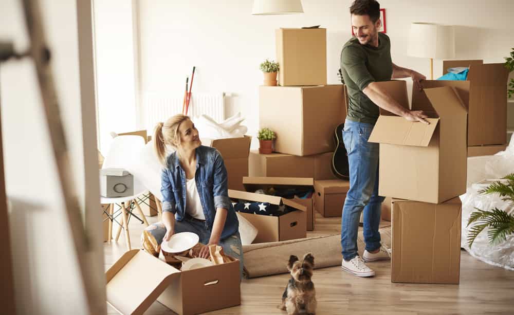 An image of two people with their dog moving boxes into their new home